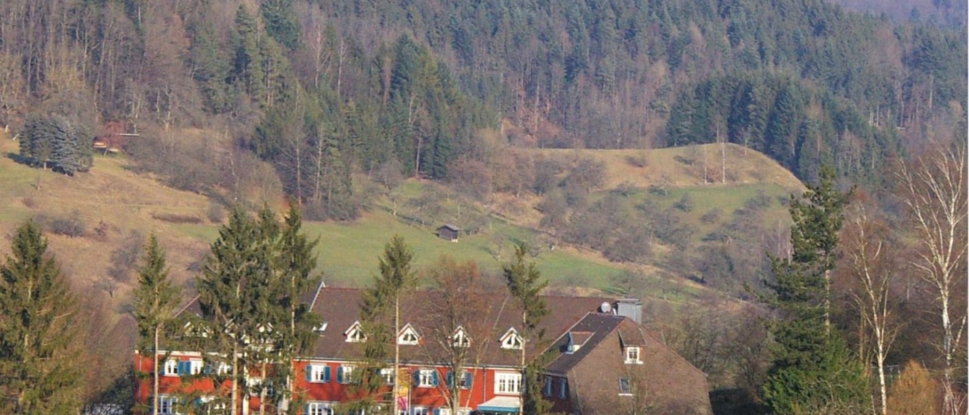 A red building with white windows stands in front of a wooded hill. Trees and bushes can be seen in the foreground., &copy; DJH Baden-W&uuml;rttemberg e.V.