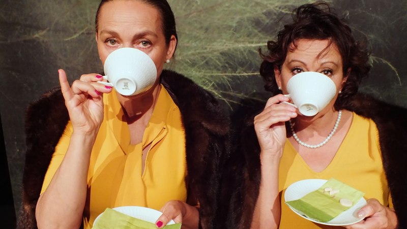 Zwei Frauen in gelber Kleidung trinken Tee aus wei&szlig;en Tassen. Sie halten Untertassen mit gr&uuml;nen Servietten und Keksen., &copy; Dieter Nelle