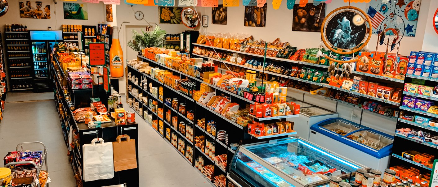Ein kleiner Supermarkt mit bunten Papierdekorationen an der Decke. Regale sind voll mit verschiedenen Lebensmitteln und Produkten., © Feinkost Latino Ventura