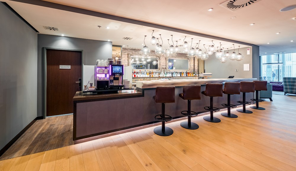 Moderne Bar mit eleganter Theke, fünf Barhockern und stilvollen Kugelleuchten. Im Hintergrund sind Flaschen und Gläser zu sehen., © Premier Inn Moderne Bar mit eleganter Theke, fünf Barhockern und stilvollen Kugelleuchten. Im Hintergrund sind Flaschen und Gläser zu sehen., © Premier Inn
