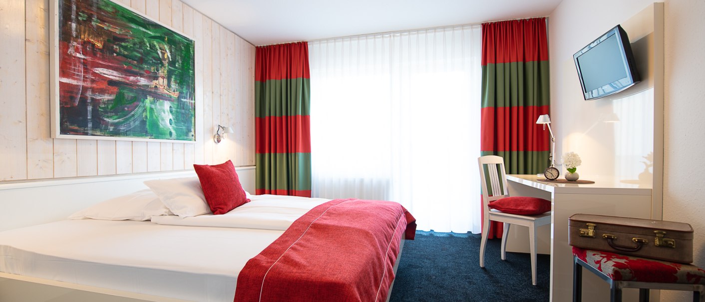 Cozy room with double bed, red bed linen, desk, chair and painting. Curtains in red and green, TV on the wall, suitcase on stool., © Hotel Weinstadt