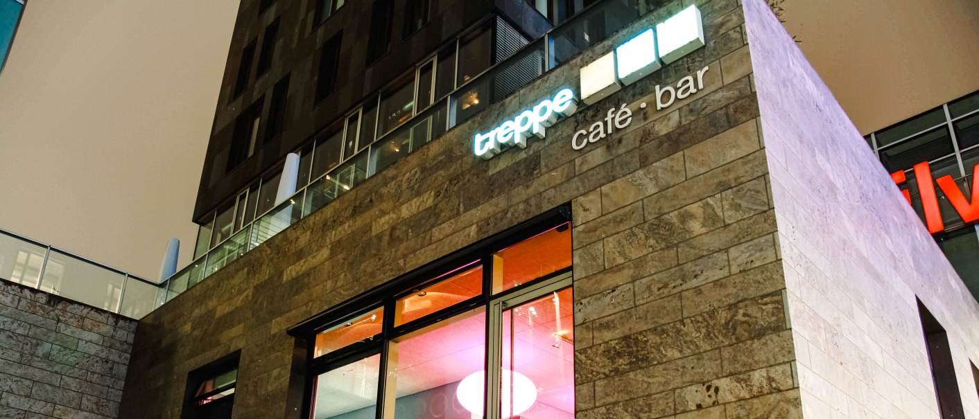 Modernes Café mit Glasfront, beleuchtetem Innenraum und Schriftzug "treppe café • bar" an der Fassade., © Café Treppe Modernes Café mit Glasfront, beleuchtetem Innenraum und Schriftzug "treppe café • bar" an der Fassade., © Café Treppe