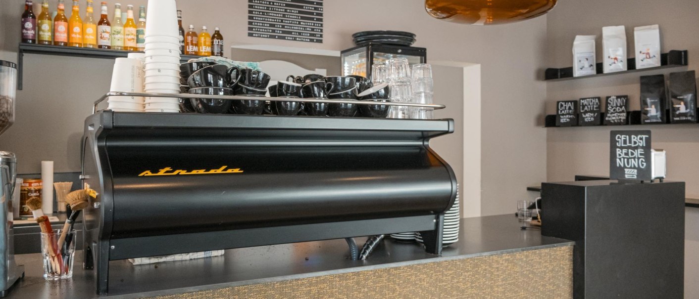 Espressomaschine in einem Café mit schwarzen Tassen, Getränkeflaschen im Hintergrund und einem Schild 'Selbstbedienung'., © SMG, Sarah Schmid