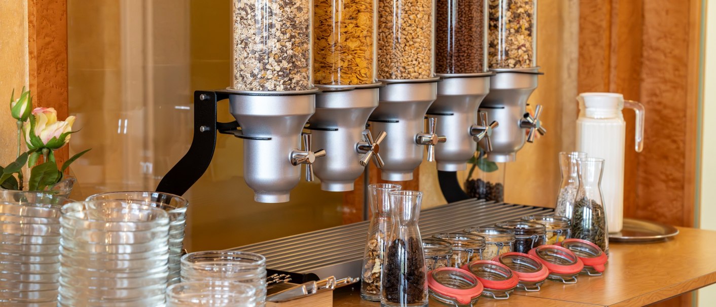Frühstücksbuffet mit fünf Müsli-Spendern, Gläsern, Karaffen und kleinen Vorratsgläsern auf einem Holztisch., © PLAZA INN Rieker Stuttgart Hauptbahnhof Frühstücksbuffet mit fünf Müsli-Spendern, Gläsern, Karaffen und kleinen Vorratsgläsern auf einem Holztisch., © PLAZA INN Rieker Stuttgart Hauptbahnhof