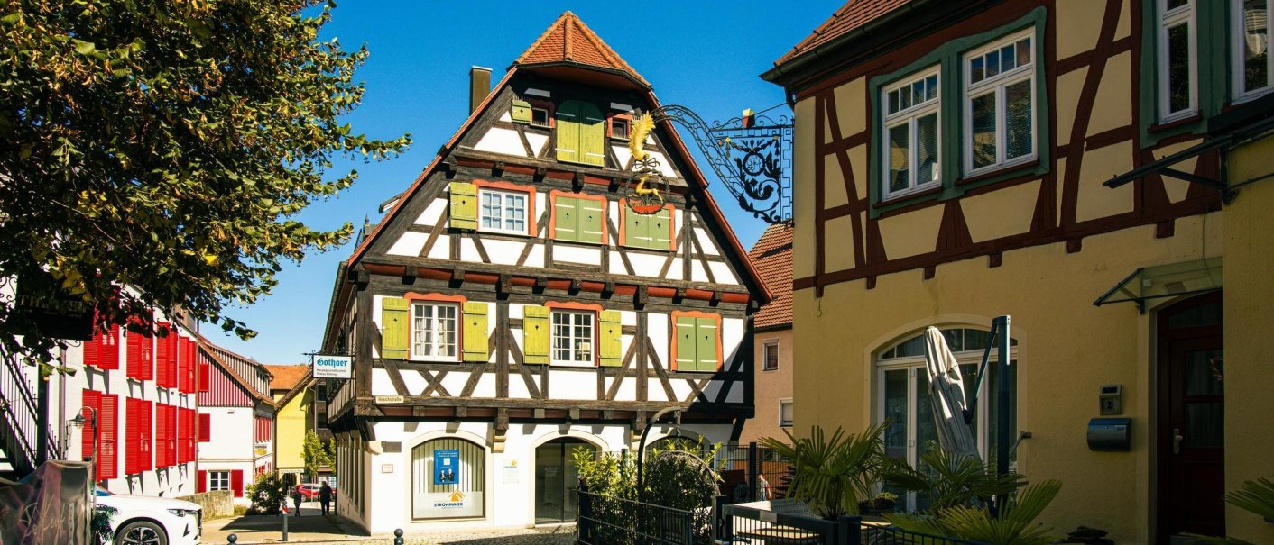 Half-timbered house in Gaildorf with green shutters and red roof, surrounded by other historic buildings and a tree in the foreground., © Stuttgart-Marketing GmbH, Sarah Schmid