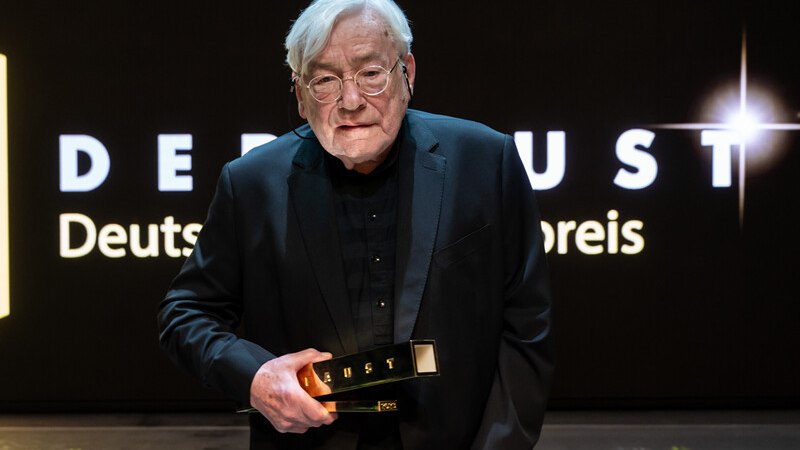 Ein älterer Mann in einem Anzug hält eine Trophäe vor einem Schild mit der Aufschrift 'DEUST Deutscher Preis'., © Württembergische Staatstheater Stuttgart Ein älterer Mann in einem Anzug hält eine Trophäe vor einem Schild mit der Aufschrift 'DEUST Deutscher Preis'., © Württembergische Staatstheater Stuttgart