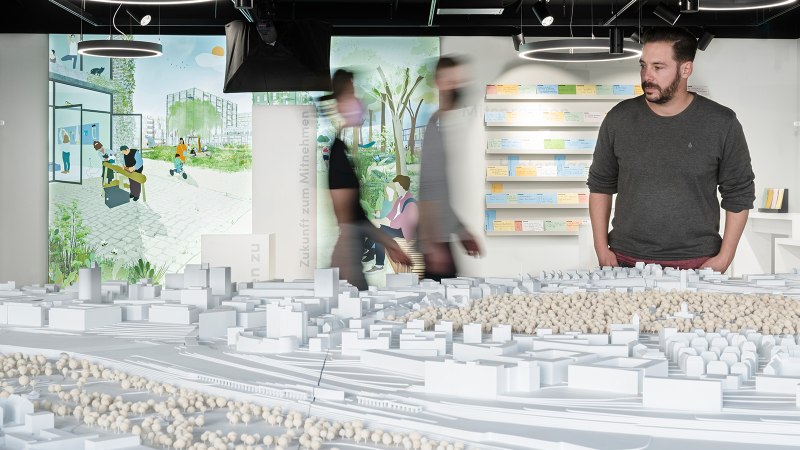 A man looks at an architectural model in an exhibition. Illustrations and notes can be seen on the wall in the background., © Dominique Brewing