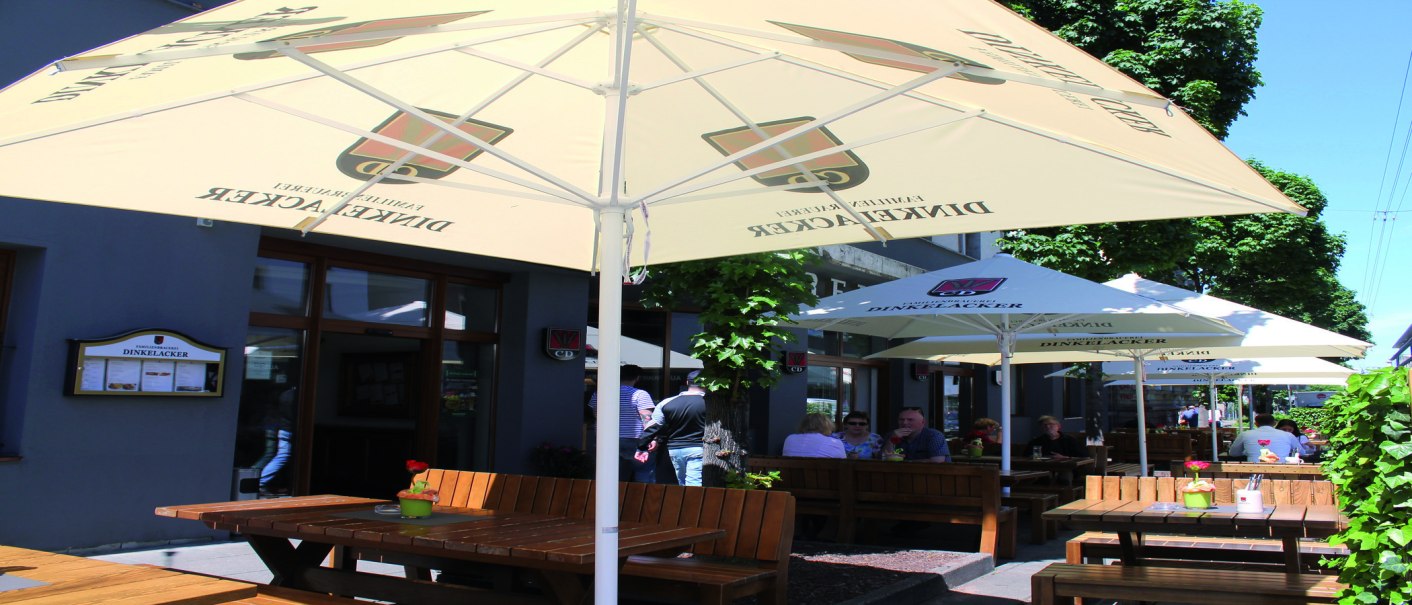 Outdoor area of the Dinkelacker brewery restaurant with wooden benches and parasols. Guests sit in the shade and enjoy the weather., © © Stuttgart-Marketing GmbH (SMG)