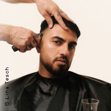 A man with a beard gets a haircut. Two hands cut his hair. He wears a black cape and looks seriously into the camera., &copy; links im Bild