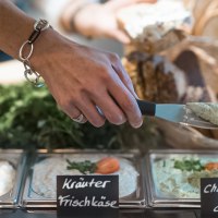 Eine Hand mit Armband nimmt Champignoncreme aus einer Schale. Daneben stehen Schalen mit Kräuterfrischkäse und Paprikacreme, im Hintergrund Brot., © Café Justus, Stuttgart
