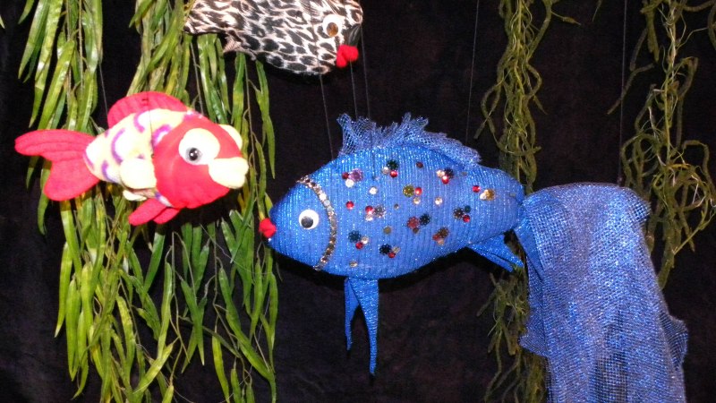 Drei bunte Stofffische, darunter ein blauer mit Glitzer, h&auml;ngen vor einem dunklen Hintergrund mit gr&uuml;nen Pflanzen., &copy; Nellys Puppentheater