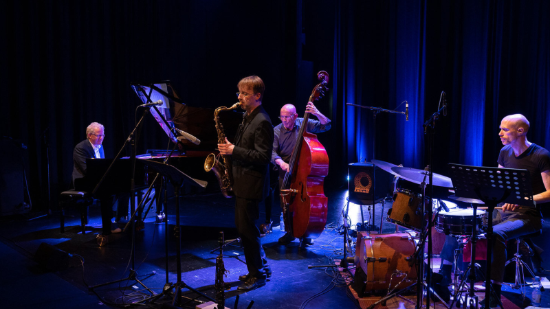 Eine Jazzband spielt auf einer Bühne. Ein Saxophonist, Pianist, Kontrabassist und Schlagzeuger sind in blauem Licht zu sehen., © Bettina Meister Eine Jazzband spielt auf einer Bühne. Ein Saxophonist, Pianist, Kontrabassist und Schlagzeuger sind in blauem Licht zu sehen., © Bettina Meister