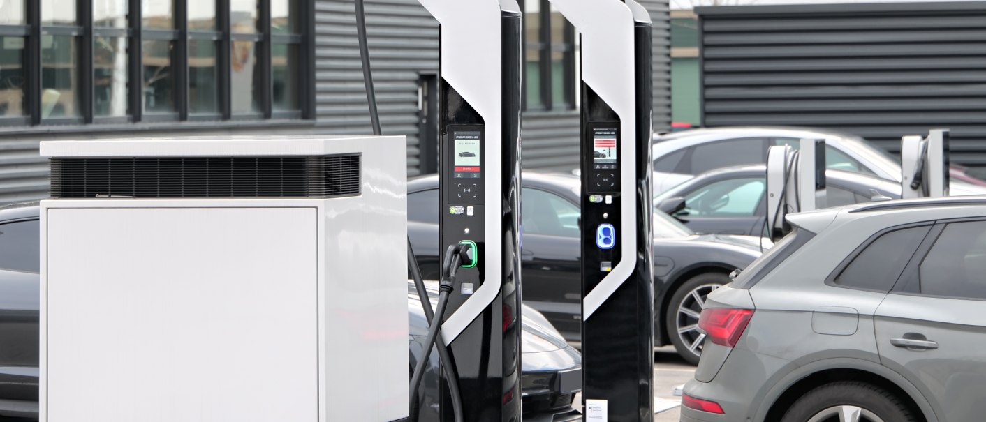 Zwei moderne Elektroladestationen auf einem Parkplatz, umgeben von geparkten Autos. Im Hintergrund ein Gebäude mit großen Fenstern., © V8 HOTEL Motorworld Region Stuttgart