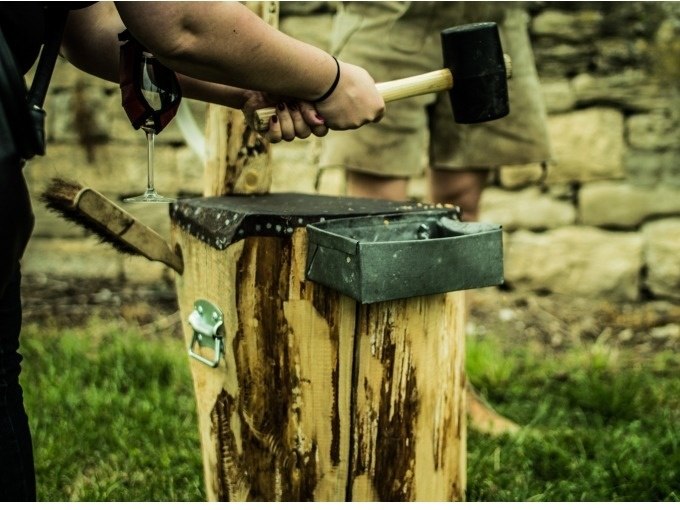 Person mit Hammer und Weinglas steht vor einem Holzblock mit Metallbehälter, im Freien auf Gras., © Cool-Tours StattReisen Person mit Hammer und Weinglas steht vor einem Holzblock mit Metallbehälter, im Freien auf Gras., © Cool-Tours StattReisen