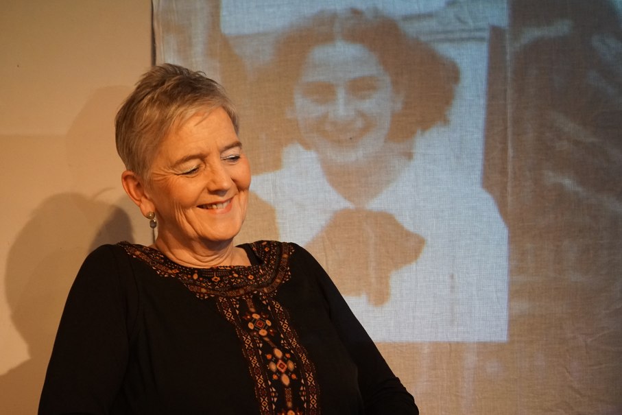 A woman smiles while a black and white photo of another woman is projected in the background. The scene is nostalgic and warm., &copy; Katja Ritter