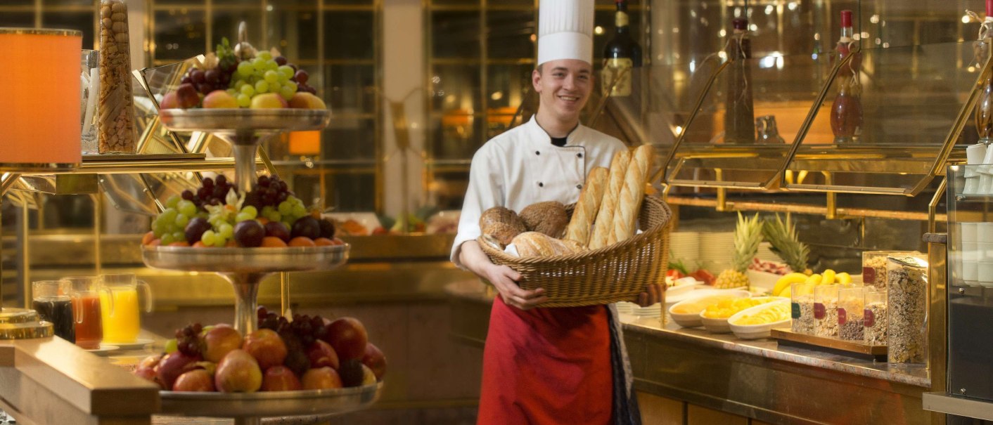 Ein Koch in Uniform hält einen Korb mit Brot vor einem reichhaltigen Frühstücksbuffet mit Obst, Saft und Müsli., © Maritim Hotelgesellschaft mbH Ein Koch in Uniform hält einen Korb mit Brot vor einem reichhaltigen Frühstücksbuffet mit Obst, Saft und Müsli., © Maritim Hotelgesellschaft mbH