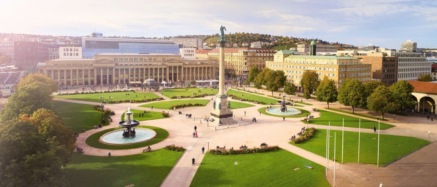 Der Schlossplatz in Stuttgart mit der Jubiläumssäule, umgeben von Grünflächen, Brunnen und historischen Gebäuden bei sonnigem Wetter., © Jean-Claude Winkler Der Schlossplatz in Stuttgart mit der Jubiläumssäule, umgeben von Grünflächen, Brunnen und historischen Gebäuden bei sonnigem Wetter., © Jean-Claude Winkler