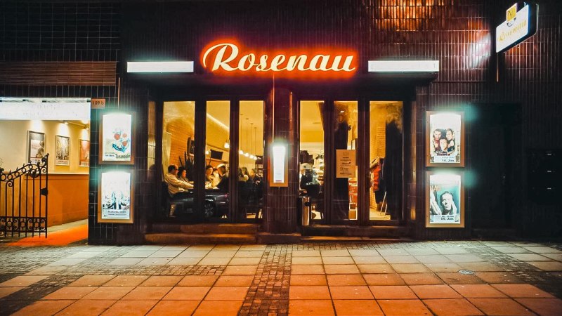 Eingang eines Lokals namens Rosenau mit leuchtendem Schild. Menschen sitzen drinnen, Plakate an den W&auml;nden, gem&uuml;tliche Atmosph&auml;re., &copy; Rosenau e. V., Stuttgart