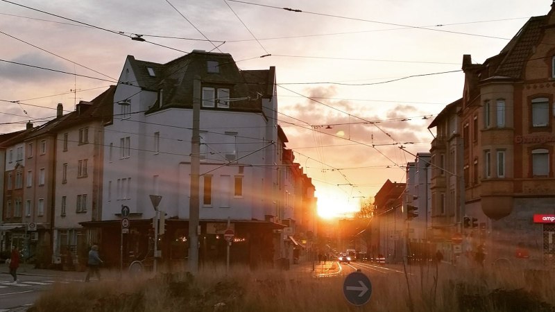 Sonnenuntergang in einer Stadtstra&szlig;e mit historischen Geb&auml;uden und Stra&szlig;enbahnkabeln. Menschen &uuml;berqueren die Stra&szlig;e, w&auml;hrend die Sonne zwischen den H&auml;usern untergeht., &copy; SMG