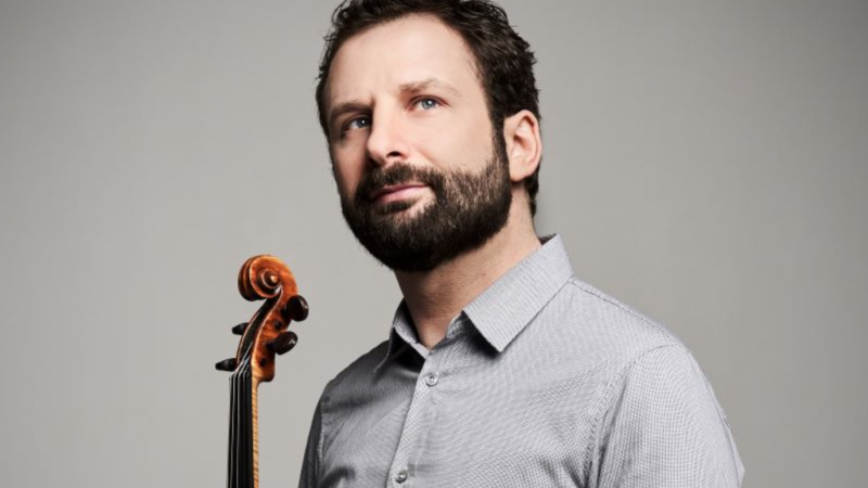 Ein Mann mit Bart hält eine Geige und blickt nachdenklich zur Seite. Er trägt ein graues Hemd vor einem neutralen Hintergrund., © Kaupo Kikkas