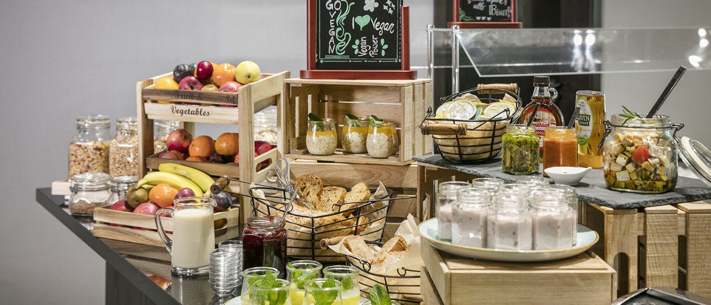 Ein Frühstücksbuffet mit Obst, Joghurt, Brot, Aufstrichen und Getränken. Ein Schild weist auf eine vegane Ecke hin., © Arcotel Hotels Ein Frühstücksbuffet mit Obst, Joghurt, Brot, Aufstrichen und Getränken. Ein Schild weist auf eine vegane Ecke hin., © Arcotel Hotels