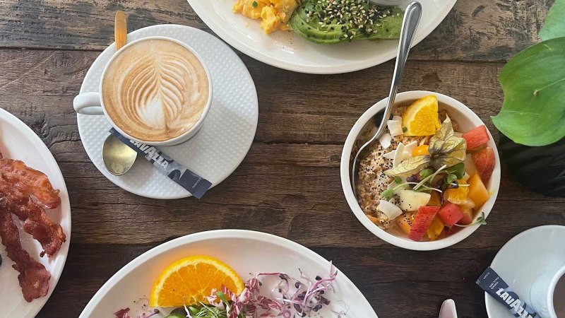 Ein reich gedeckter Frühstückstisch mit Kaffee, Pancakes mit Speck, Avocado mit Rührei und einer Schale mit frischem Obst., © SMG