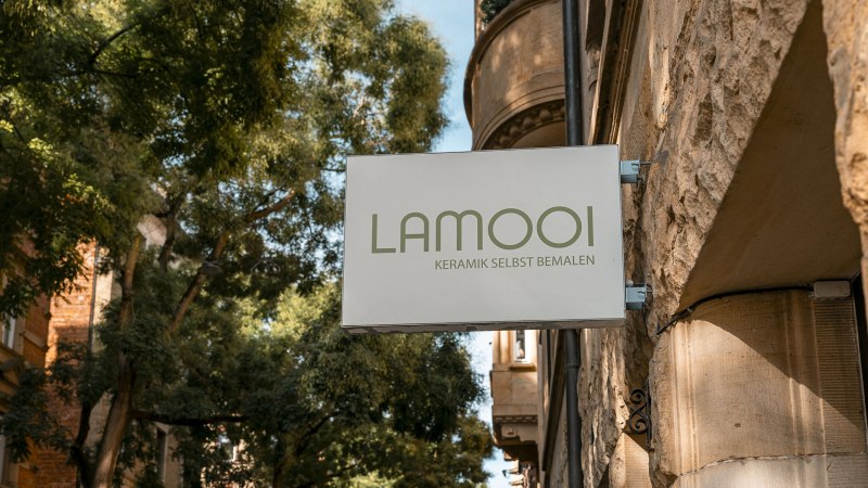 Schild mit 'LAMOOI Keramik selbst bemalen' an einer Steinwand, umgeben von B&auml;umen., &copy; SMG, Sarah Schmid