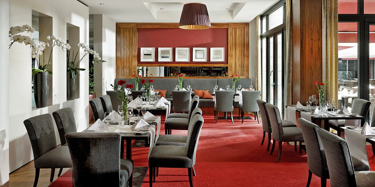 Elegant restaurant with gray chairs, red carpet and white tablecloths. Large windows and orchids decorate the room., © Parkhotel Stuttgart Messe-Airport Elegant restaurant with gray chairs, red carpet and white tablecloths. Large windows and orchids decorate the room., © Parkhotel Stuttgart Messe-Airport