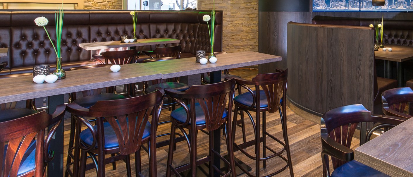 Moderner Pub mit Holztischen, Barhockern und dekorativen Vasen. Ein grünes Neonschild an der Wand zeigt "Mike's urban pub"., © Ibis Styles Stuttgart Moderner Pub mit Holztischen, Barhockern und dekorativen Vasen. Ein grünes Neonschild an der Wand zeigt "Mike's urban pub"., © Ibis Styles Stuttgart