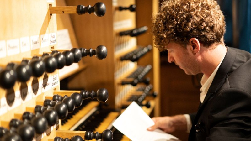 Ein Mann in einem Anzug spielt an einer Orgel und hält ein Blatt Papier in der Hand. Die Orgelregister sind deutlich sichtbar., © Johannes Schaugg Ein Mann in einem Anzug spielt an einer Orgel und hält ein Blatt Papier in der Hand. Die Orgelregister sind deutlich sichtbar., © Johannes Schaugg