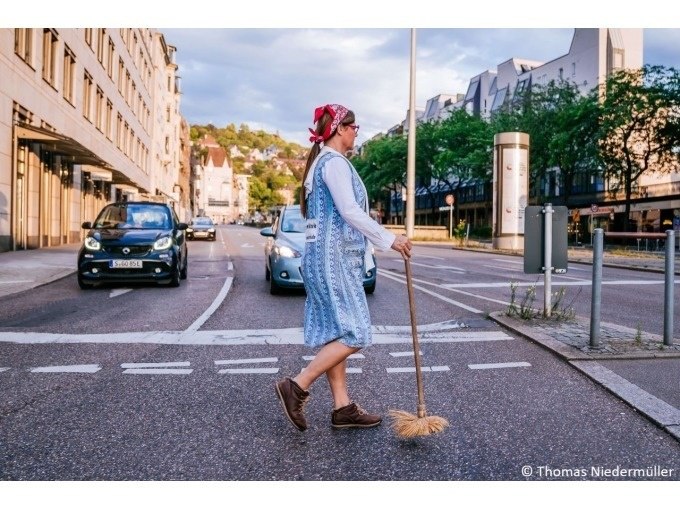 Eine Frau in traditioneller Kleidung überquert eine Straße mit einem Besen. Im Hintergrund sind Autos und Gebäude zu sehen., © Stuttgart Marketing GmbH