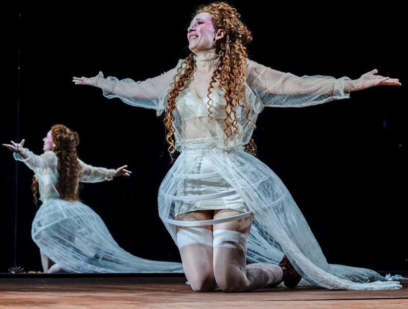 Eine Frau mit lockigem Haar kniet in einem weißen, transparenten Kleid mit ausgebreiteten Armen vor einem Spiegel., © Württembergische Staatstheater Stuttgart