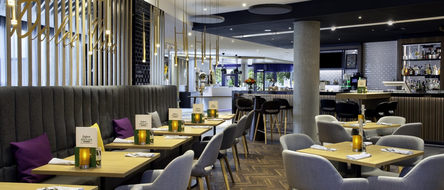 Modern restaurant with elegant furnishings, wooden tables, gray chairs and stylish lighting. A bar with a bottle rack in the background., © Holiday Inn Stuttgart Modern restaurant with elegant furnishings, wooden tables, gray chairs and stylish lighting. A bar with a bottle rack in the background., © Holiday Inn Stuttgart