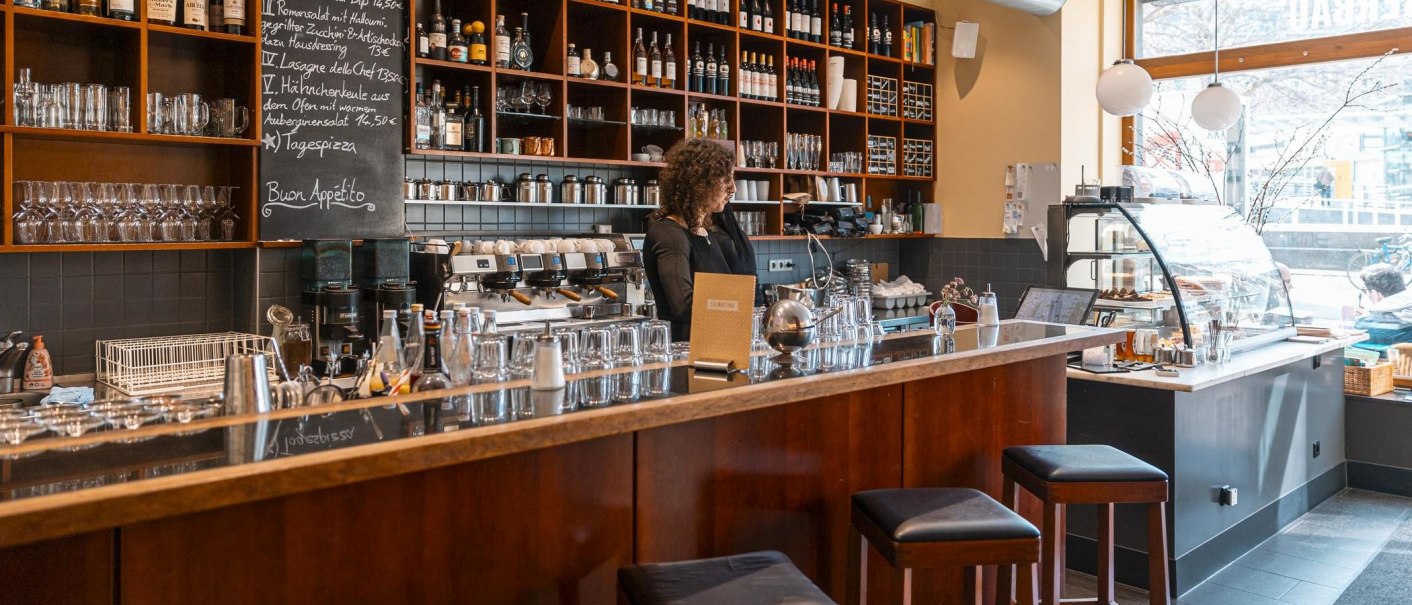 Ein gemütliches Café mit einer langen Theke, Regalen voller Flaschen und Gläser. Eine Person steht hinter der Theke, es gibt Sitzgelegenheiten und eine Vitrine., © SMG, Sarah Schmid