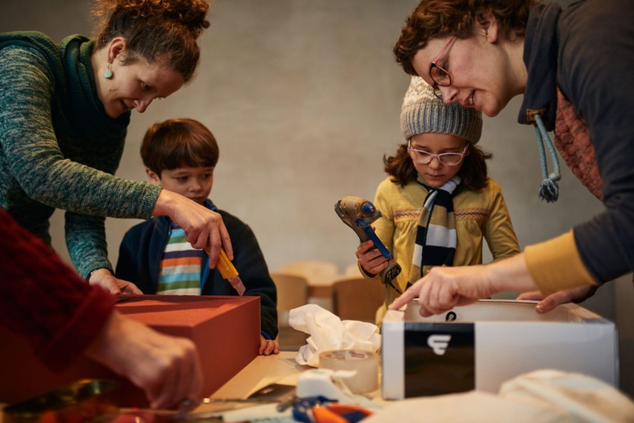 Zwei Erwachsene und zwei Kinder basteln an einem Tisch mit Kartons und Werkzeugen. Die Kinder halten Klebepistolen., &copy; Alex Wunsch