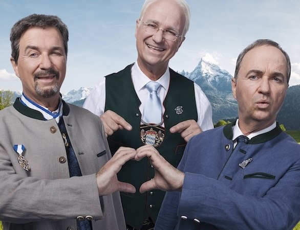 Drei Männer in bayerischer Tracht stehen vor einer Bergkulisse. Zwei formen mit ihren Händen ein Herz, während der dritte lächelnd in der Mitte steht., © Renitenztheater Stuttgart e.V.