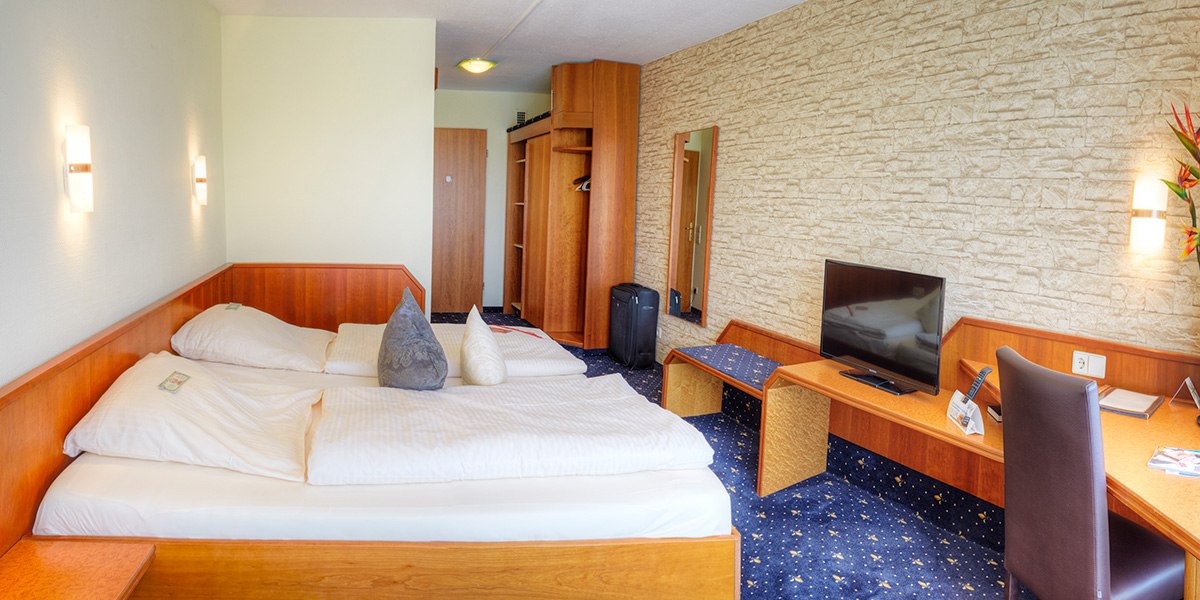 A cozy double room with wooden furniture, double bed, desk, TV and suitcase. The walls are partly designed with a stone look., © Kemnater Hof GmbH