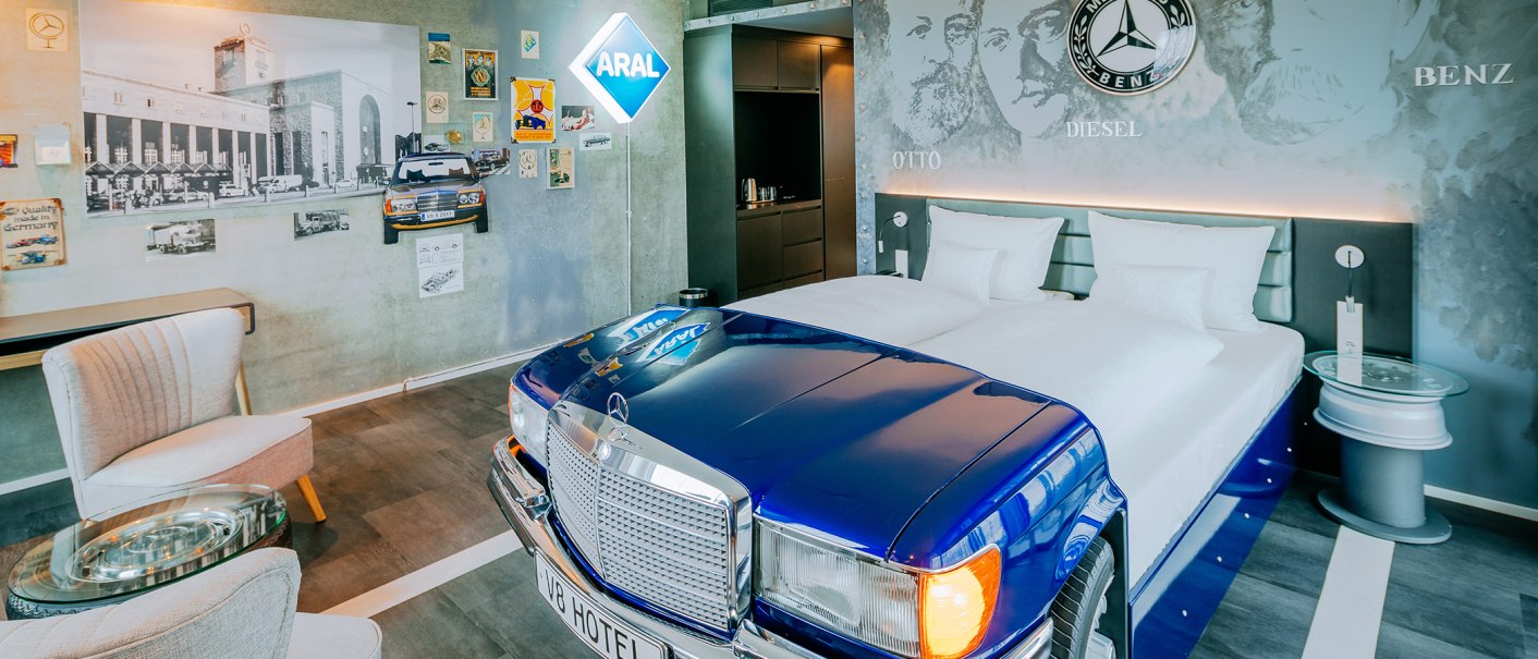 Ein Hotelzimmer im V8 Hotel mit einem Bett in Form eines blauen Mercedes-Autos, Wanddekorationen und einem ARAL-Schild., © Thomas Niedermüller Ein Hotelzimmer im V8 Hotel mit einem Bett in Form eines blauen Mercedes-Autos, Wanddekorationen und einem ARAL-Schild., © Thomas Niedermüller