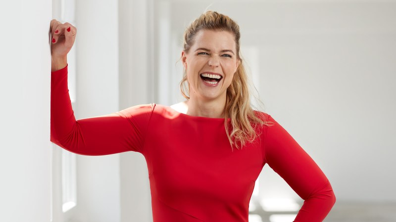 Eine Frau in einem roten Kleid lacht herzlich und lehnt sich an eine weiße Wand in einem hellen Raum., © club traube