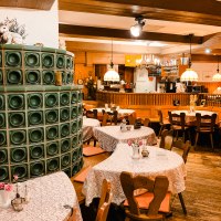 Gemütliches Restaurant mit Holzmöbeln, Tischdecken und einem grünen Kachelofen. Lampen hängen von der Decke, Bar im Hintergrund., © Weinstube Kachelofen Gemütliches Restaurant mit Holzmöbeln, Tischdecken und einem grünen Kachelofen. Lampen hängen von der Decke, Bar im Hintergrund., © Weinstube Kachelofen