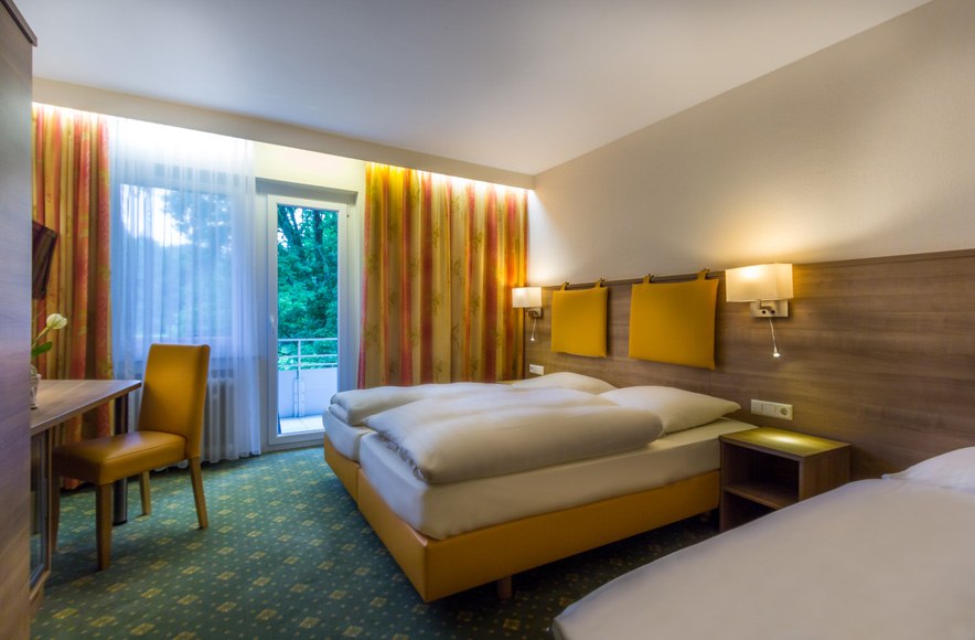 A modern hotel room with two beds, a desk, yellow accents, and a balcony. Large windows with curtains, carpeted floors, and wall sconces., &copy; Kulinarium an der Glems