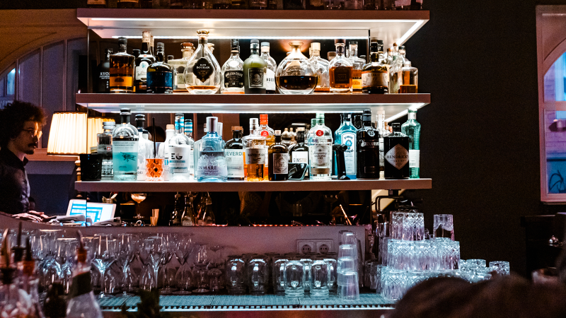 Bar mit beleuchteten Regalen voller Spirituosenflaschen, Gläsern und einem Barkeeper im Hintergrund. Gemütliche Atmosphäre mit gedämpftem Licht., © SMG, Sarah Schmid Bar mit beleuchteten Regalen voller Spirituosenflaschen, Gläsern und einem Barkeeper im Hintergrund. Gemütliche Atmosphäre mit gedämpftem Licht., © SMG, Sarah Schmid