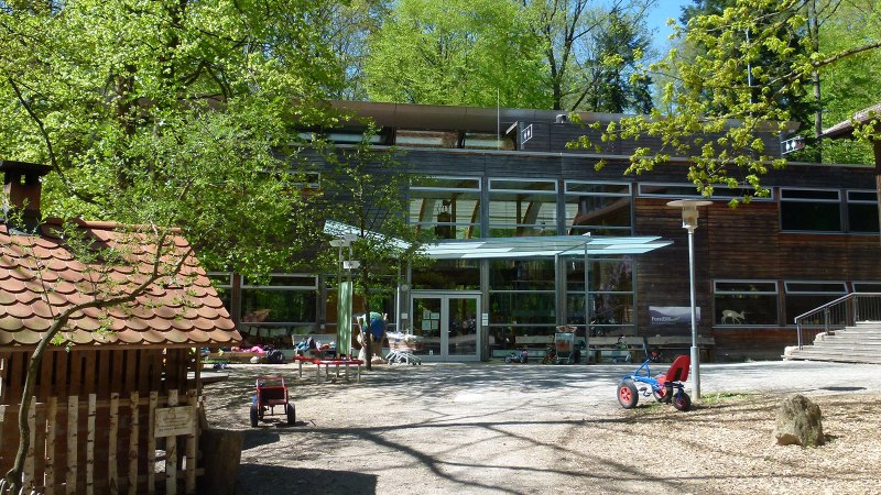 Inner courtyard of the House of the Forest with toy vehicles, trees and a small wooden house. Modern glass façade in the background., © Haus des Waldes Inner courtyard of the House of the Forest with toy vehicles, trees and a small wooden house. Modern glass façade in the background., © Haus des Waldes