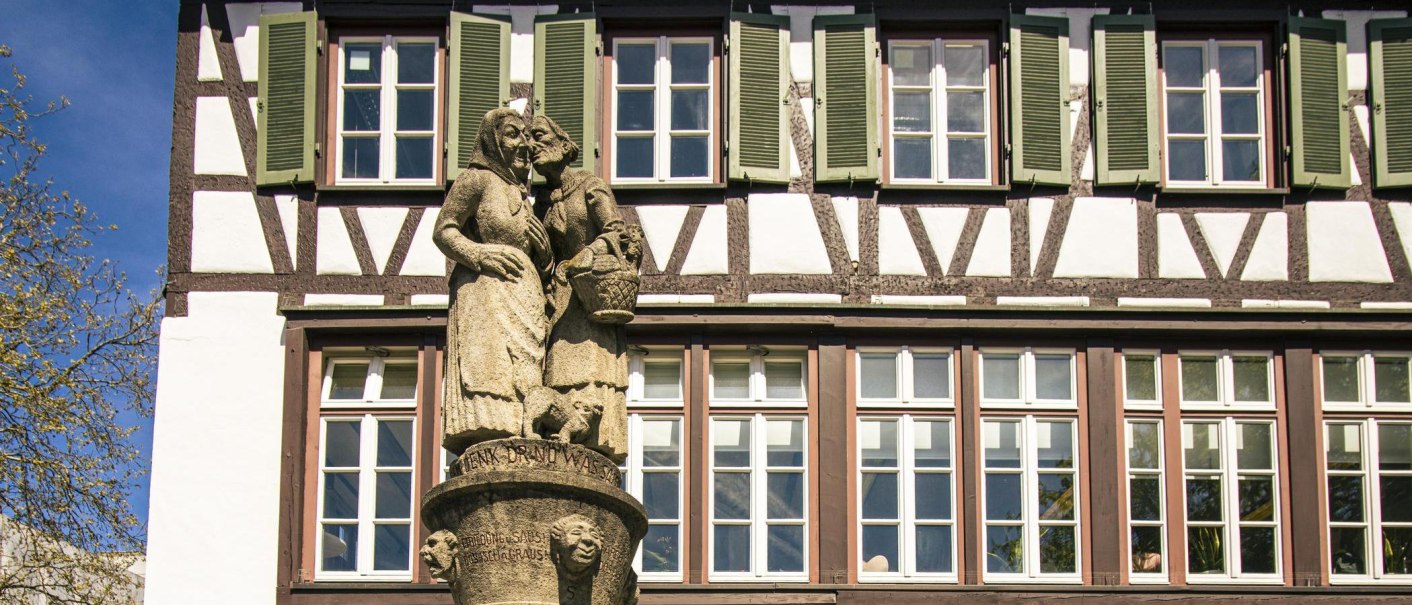 The "Schwätzweiberbrunnen" fountain in Sindelfingen features two stone figures of women engaged in conversation. A half-timbered house can be seen in the background., © SMG Stuttgart Marketing GmbH - Sarah Schmid The "Schwätzweiberbrunnen" fountain in Sindelfingen features two stone figures of women engaged in conversation. A half-timbered house can be seen in the background., © SMG Stuttgart Marketing GmbH - Sarah Schmid