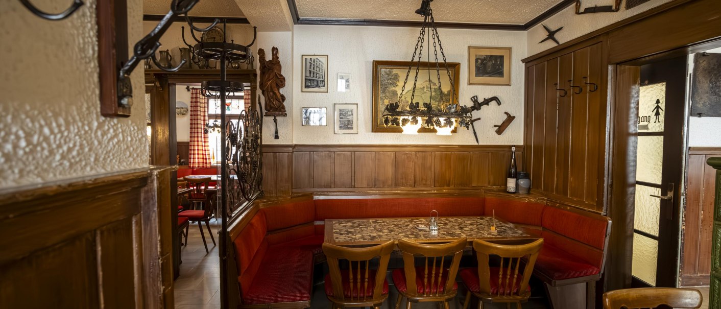 Gemütliche Weinstube mit Holzmöbeln, roten Polstern und rustikaler Dekoration. An den Wänden hängen Bilder und Werkzeuge, ein Kronleuchter beleuchtet den Raum., © Stuttgart Marketing GmbH, Sarah Schmid Gemütliche Weinstube mit Holzmöbeln, roten Polstern und rustikaler Dekoration. An den Wänden hängen Bilder und Werkzeuge, ein Kronleuchter beleuchtet den Raum., © Stuttgart Marketing GmbH, Sarah Schmid