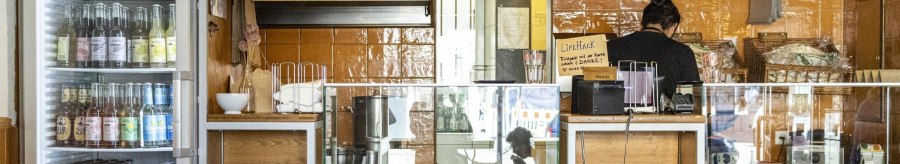 A snack bar with drinks fridge, counter and menu board. One person works behind the counter. The walls are tiled in orange., © SMG, Sarah Schmid