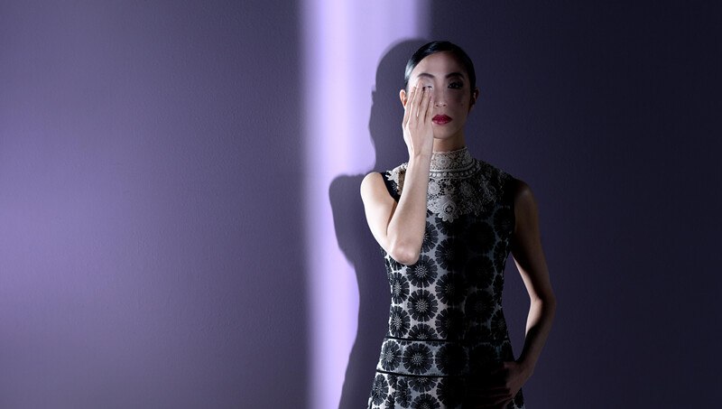 Frau in elegantem Kleid bedeckt ein Auge mit der Hand, steht vor lila beleuchteter Wand., © Württembergische Staatstheater Stuttgart Frau in elegantem Kleid bedeckt ein Auge mit der Hand, steht vor lila beleuchteter Wand., © Württembergische Staatstheater Stuttgart