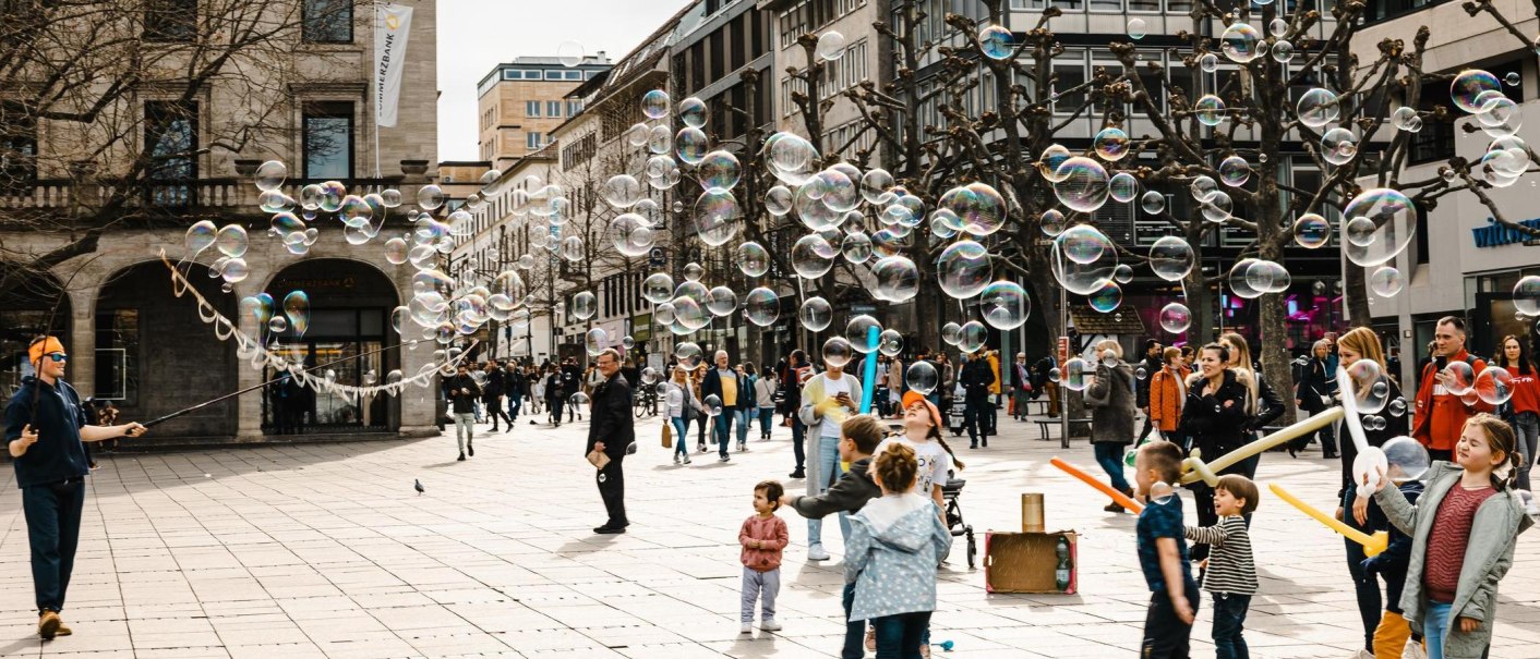 Ein Stra&szlig;enk&uuml;nstler erzeugt Seifenblasen auf einer belebten Stra&szlig;e. Kinder spielen begeistert mit den Blasen, w&auml;hrend Passanten zuschauen., &copy; Stuttgart-Marketing GmbH, Sarah Schmid