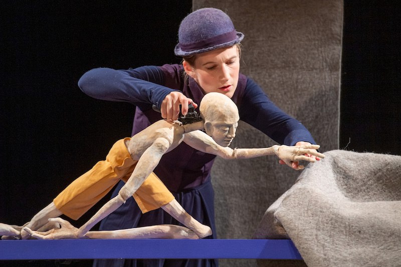 Eine Puppenspielerin mit Hut manipuliert eine bewegliche Puppe auf einer Bühne. Die Puppe trägt gelbe Shorts und ist in einer dynamischen Pose., © FITZ Das Theater animierter Formen