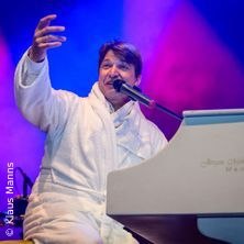 Ein Mann im weißen Bademantel sitzt an einem weißen Klavier auf einer Bühne und singt ins Mikrofon. Der Hintergrund ist bunt beleuchtet., © links im Bild